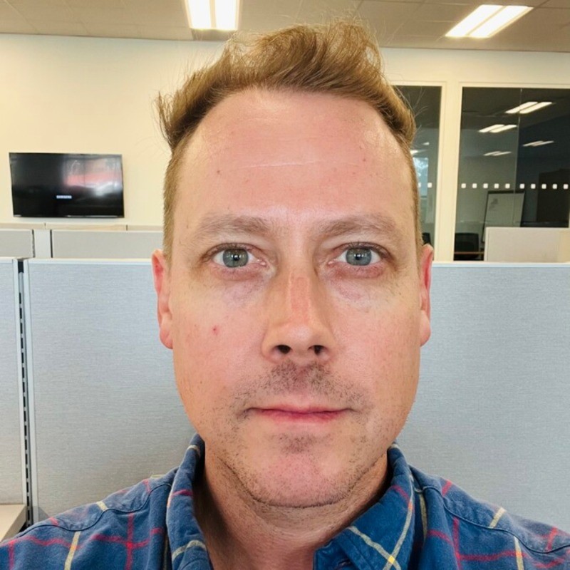 Close-up portrait of a man with blue eyes and light brown hair, wearing a blue plaid shirt in a gray office cubicle background.