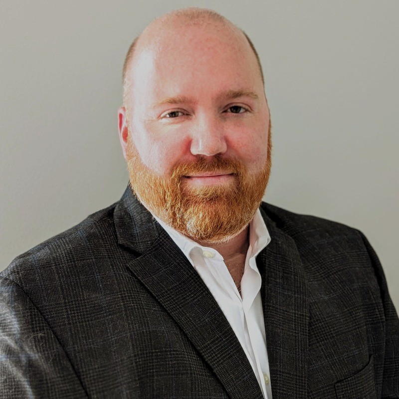 Head-and-shoulders portrait of a man with a red beard wearing a dark checkered blazer and white shirt, smiling softly against a gray background.