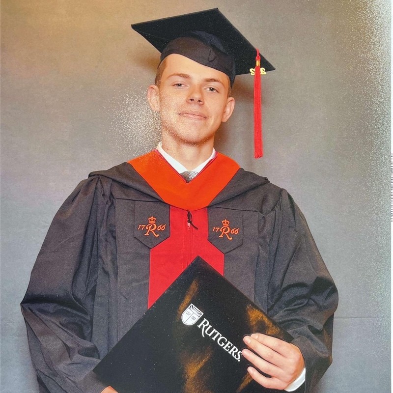 Graduate in cap and gown holding a Rutgers diploma cover, posing for photo.