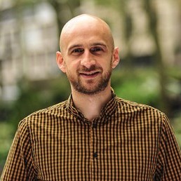 Man with a shaved head and short beard, wearing a brown checkered shirt, standing outdoors and smiling in a park-like setting.