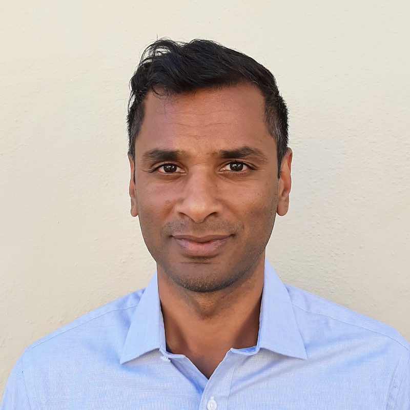 Portrait of a man with short dark hair wearing a light blue button-down shirt, against a beige wall.