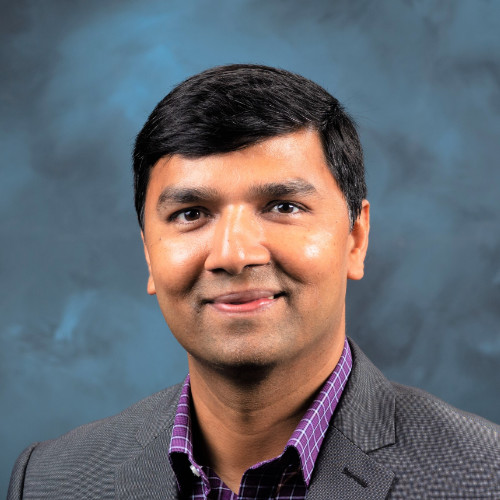 Professional headshot of a smiling man in a gray blazer against a blue studio backdrop.