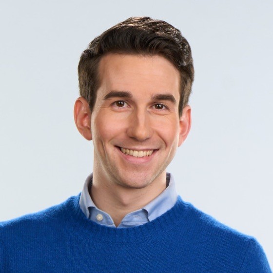 Portrait of a smiling man with short dark hair wearing a blue sweater against a light background.