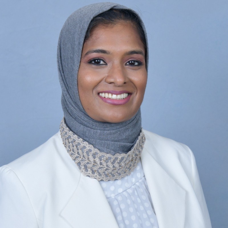 Smiling woman wearing a gray hijab and white blazer against a blue background with a lace collar detail.